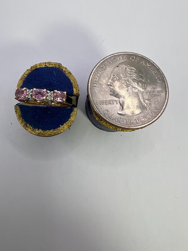 Puede incluir: Un anillo de oro con tres piedras preciosas rosas de corte ovalado y dos peque&ntilde;as piedras transparentes, exhibido en un soporte de terciopelo azul junto a una moneda de 25 centavos de d&oacute;lar para mostrar la escala de la joya.