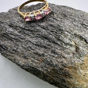 Puede incluir: Un anillo de oro con tres piedras preciosas rosas de corte ovalado dispuestas horizontalmente, separadas por peque&ntilde;as piedras transparentes engastadas en una base de metal plateado. La banda es un aro de oro sencillo y liso.