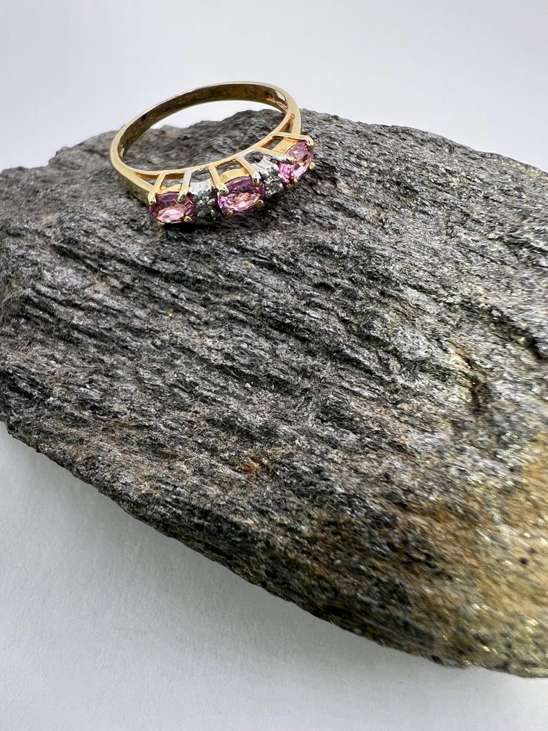 Puede incluir: Un anillo de oro con tres piedras preciosas rosas de corte ovalado dispuestas horizontalmente, separadas por peque&ntilde;as piedras transparentes engastadas en una base de metal plateado. La banda es un aro de oro sencillo y liso.