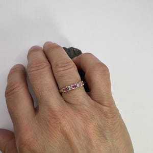 Puede incluir: Un anillo de banda dorada con tres piedras preciosas rosas redondas que se alternan con dos piedras peque&ntilde;as transparentes en un engaste de garras. El anillo se lleva en un dedo, mostrando una fila horizontal de cinco piedras en una banda.