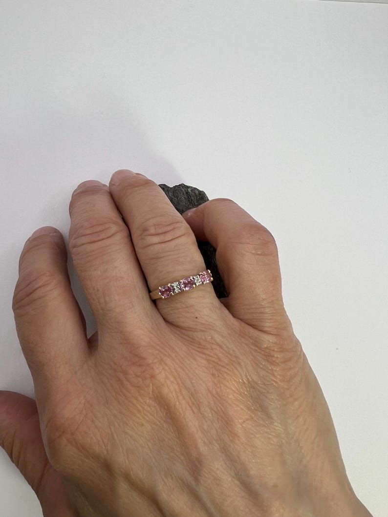Puede incluir: Un anillo de banda dorada con tres piedras preciosas rosas redondas que se alternan con dos piedras peque&ntilde;as transparentes en un engaste de garras. El anillo se lleva en un dedo, mostrando una fila horizontal de cinco piedras en una banda.