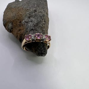 Puede incluir: Un anillo de oro con tres piedras preciosas rosas redondas en fila, acentuadas por peque&ntilde;as piedras claras entre ellas. El anillo est&aacute; colocado alrededor de un trozo de roca natural gris oscuro y dentado, mostrando el acabado met&aacute;lico pulido.