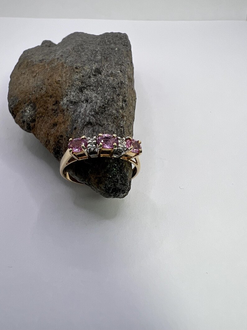 Puede incluir: Un anillo de oro con tres piedras preciosas rosas redondas en fila, acentuadas por peque&ntilde;as piedras claras entre ellas. El anillo est&aacute; colocado alrededor de un trozo de roca natural gris oscuro y dentado, mostrando el acabado met&aacute;lico pulido.