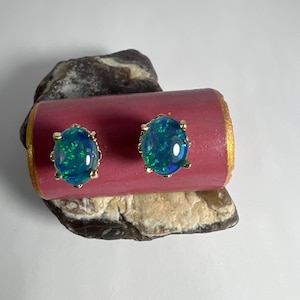 May include: A pair of gold stud earrings, each featuring an oval-shaped opal with vibrant blue and green hues. The opals are set in a gold bezel setting. The earrings are displayed on a pink and gold surface, resting on a rock.