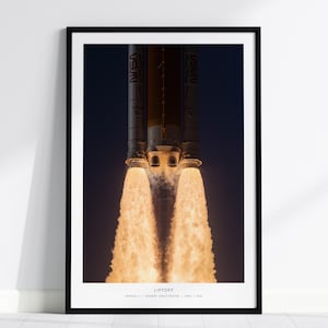 Puede incluir: Una impresión artística enmarcada que presenta una fotografía vertical del lanzamiento de un cohete por la noche. El cohete muestra el logotipo de la NASA en el lateral, con llamas de color naranja brillante saliendo de los motores. El texto inferior dice: LIFTOFF, ARTEMIS I, KENNEDY SPACE CENTER, APRIL 2022.