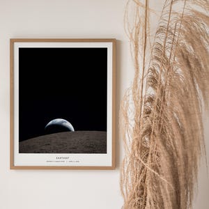 May include: A framed print of the Earth rising over the lunar surface, a classic image from the Apollo 8 mission. The print features a black sky, a gray lunar surface, and the Earth in shades of blue and white. The print is in a wooden frame.