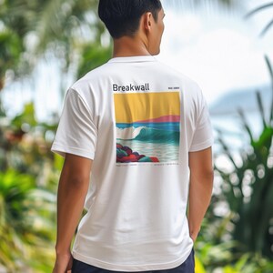 May include: White short-sleeved t-shirt with a colourful graphic on the back. The graphic depicts a wave, rocks, and the text "Breakwall". The design incorporates yellow, blue, pink, and red. The shirt is made of a soft material.
