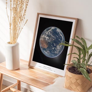 May include: A framed print of Earth from space, with the words "HELLO WORLD" at the bottom. The print is in a light wood frame and is displayed on a wooden table. A white vase with dried plants and a potted plant are also on the table.