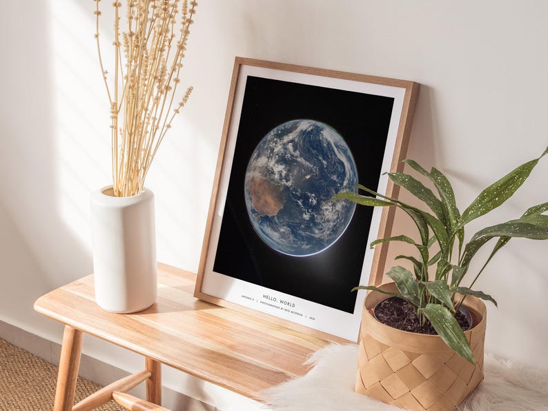 May include: A framed print of Earth from space, with the words "HELLO WORLD" at the bottom. The print is in a light wood frame and is displayed on a wooden table. A white vase with dried plants and a potted plant are also on the table.