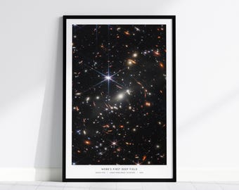 Könnte beinhalten: Gerahmter Druck des ersten Deep-Field-Bildes des James-Webb-Weltraumteleskops, das ein dichtes Feld von Galaxien und Sternen vor dunklem Hintergrund zeigt. Das Bild ist in einem schwarzen Rahmen und enthält den Text "WEBB'S FIRST DEEP FIELD".