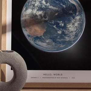 May include: Framed print of Earth from space, showcasing blue oceans, brown continents, and white clouds against a black background. The print is in a light wood frame. Text at the bottom reads "HELLO, WORLD" and details about the photograph.