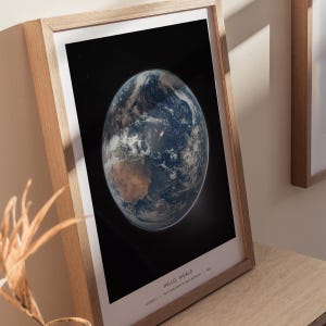 May include: A framed print featuring a detailed image of Earth, showcasing continents and cloud formations against a black space background. The print is in a light-colored wooden frame. The text "HELLO WORLD" is at the bottom.