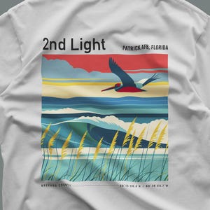 May include: A white t-shirt featuring a graphic print of a pelican flying over ocean waves and beach grass. The text reads 2nd Light, Patrick AFB, Florida, Brevard County, and coordinates 28 15 09.2 N / 80 36 09.7 W in a colorful coastal scene.