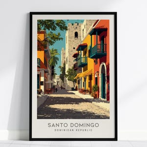 May include: A framed art print depicting a street scene in Santo Domingo, Dominican Republic. The illustration features vibrant orange and yellow colonial buildings with teal balconies, a stone tower, and a cobblestone street under a blue sky.