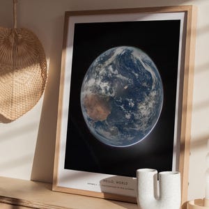 May include: A framed print of Earth from space, showcasing the planet's blue oceans, brown landmasses, and swirling white clouds against a black background. The print is in a light wood frame. Text at the bottom reads "HELLO, WORLD".