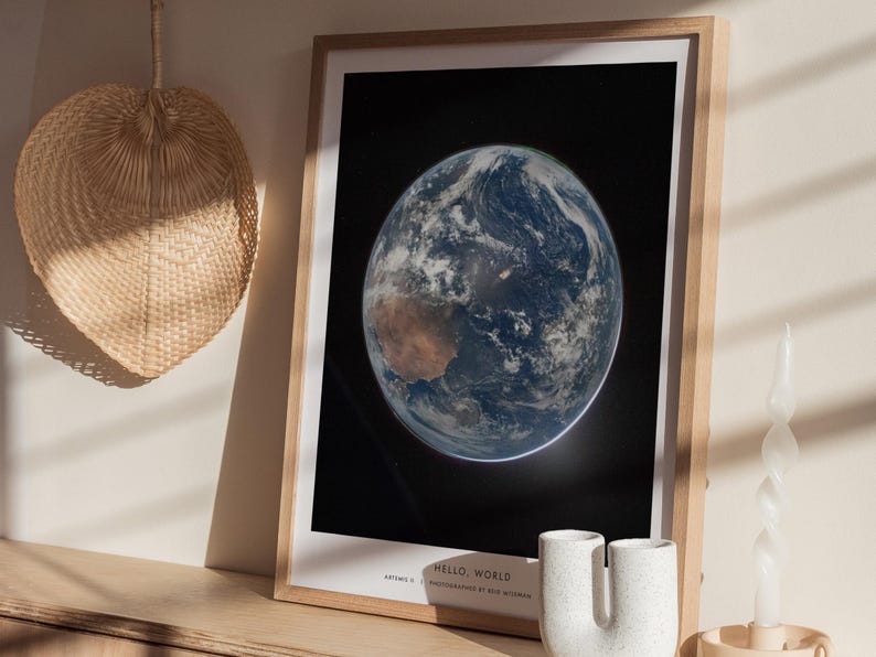 May include: A framed print of Earth from space, showcasing the planet's blue oceans, brown landmasses, and swirling white clouds against a black background. The print is in a light wood frame. Text at the bottom reads "HELLO, WORLD".