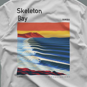 Op de afbeelding: Een wit t-shirt met een grafische print van Skeleton Bay, Namibië. Het ontwerp toont een gestileerde oceaangolf, een rode berg en een zonsondergang. De tekst bevat 'Skeleton Bay', 'NAMIBIA' en de coördinaten '22 37 12.00 S / 14 31 48.00 E'.