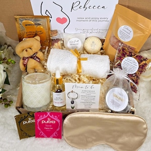May include: A gift box filled with self-care items. Includes a teddy bear, candle, tea, bath salts, rose body oil, eye mask, and a card that says "Rebecca relax and enjoy every moment of this journey."