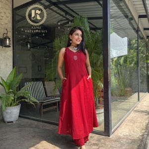 Puede incluir: Un vestido rojo sin mangas con detalles bordados y bolsillos, combinado con pantalones rojos a juego. La imagen también muestra el texto "KANU ENTERPRISES" en un logotipo circular en el cristal.