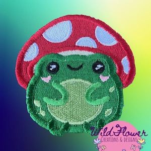 May include: An embroidered patch featuring a green frog with a light green belly, pink cheeks, and black eyes, wearing a large red mushroom cap with light blue spots. The patch includes the text Wild Flower CREATIONS & DESIGNS with a bird icon.