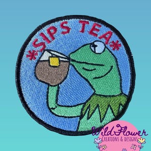 May include: A circular embroidered patch featuring a green frog character drinking from a brown mug with a yellow tea bag tag. The background is light blue with a black border. The red text reads *SIPS TEA* in an arc above the frog character.