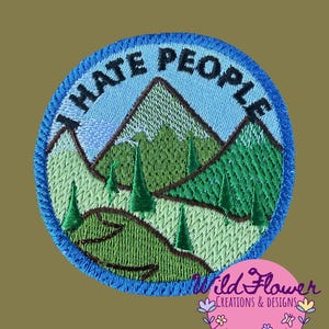May include: A circular embroidered patch featuring a mountain landscape with green hills, dark green pine trees, and a light blue sky. The text "I HATE PEOPLE" is stitched in black along the top edge of the patch, which is bordered by a thick blue thread.