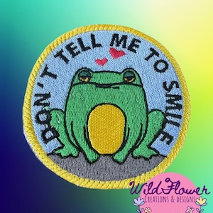 May include: A circular embroidered patch featuring a green frog with a yellow belly on a light blue background. The text DON'T TELL ME TO SMILE is stitched in black around the frog, with two small red hearts above its head and a yellow border.