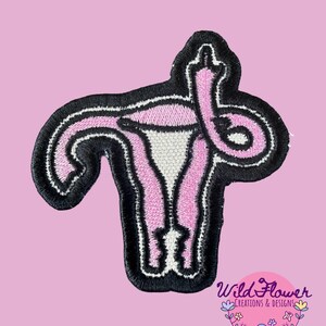 May include: An embroidered patch featuring a stylized anatomical uterus in shades of pink and white with a black border. One of the fallopian tubes is shaped like an upright middle finger. The text Wild Flower Creations & Designs is visible.