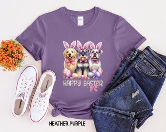 Puede incluir: Camiseta morada con un gráfico de tres perros con orejas de conejo y sosteniendo huevos de Pascua. El texto "Happy Easter" está impreso debajo de los perros.