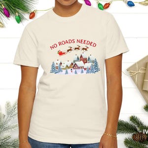 May include: Ivory t-shirt with a Christmas-themed graphic. The design shows Santa's sleigh and reindeer above a snowy village. The text "NO ROADS NEEDED" is printed in red. The shirt is a casual style, perfect for the festive season. The shirt is a comfortable, casual style.