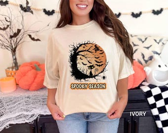 May include: Ivory-colored t-shirt with a Halloween-themed graphic. The design features a full moon, bats, a black cat, and the words "Spooky Season." The shirt is a comfortable, casual style.
