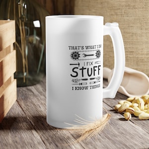 Peut inclure: Une tasse en céramique blanche givrée avec un motif noir qui dit "That's what I do I fix stuff I know things". Le motif comprend également une chaîne, une clé, un marteau et d'autres outils.
