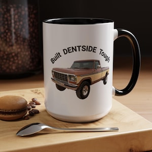 May include: White ceramic mug with a black interior and handle. The mug features a brown and tan pickup truck graphic with the text "Built DENTSIDE Tough" in a curved font.