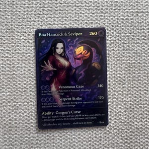 Può includere: Carta collezionabile con Boa Hancock e Seviper su sfondo viola scuro. La carta mostra i nomi degli attacchi e i valori dei danni. Il testo "All who dare defy beauty... shall turn to stone" è anch'esso presente.