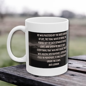 May include: White ceramic mug with a black rectangular design featuring a quote by Jack London. The quote is set against a backdrop of a mountain landscape and starry sky. The mug has a curved handle.