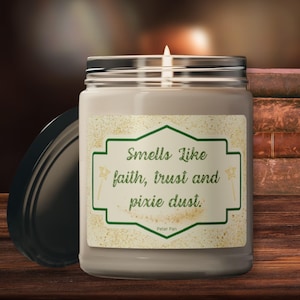 Op de afbeelding: Een bruine glazen kaars pot met een wit label waarop staat "Smells Like faith, trust and pixie dust." Het label heeft ook een groene rand en gouden glitter. De tekst "Peter Pan" staat onderaan het label.