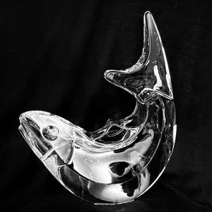 May include: A clear glass sculpture shaped like a leaping fish. The object features a smooth, reflective surface with defined contours representing the head, body, and tail fin, captured in a curved, upward-arching position against a dark background.