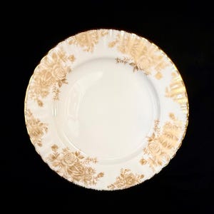 May include: A round white porcelain plate featuring a scalloped edge with a gold trim. The rim is decorated with a repeating pattern of light brown floral rose motifs and foliage, leaving the center of the plate plain and white.