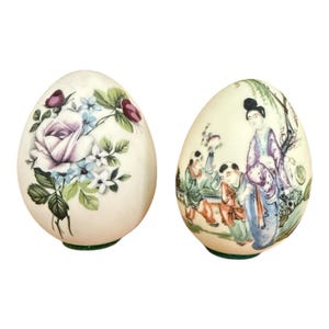 May include: Two egg-shaped decorative objects with smooth surfaces. One egg features a hand-painted floral design with a large purple rose and small blue flowers. The second egg displays a painted scene of three figures in traditional attire.