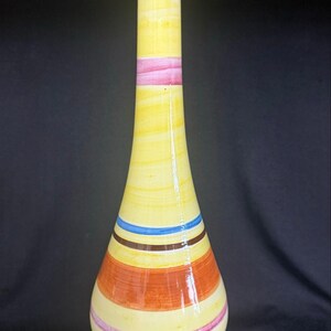 May include: A tall, slender ceramic vase with a glossy finish, featuring a pale yellow base color decorated with horizontal bands in shades of orange, pink, brown, and blue. The vase has a narrow neck that widens toward a rounded, stable base.