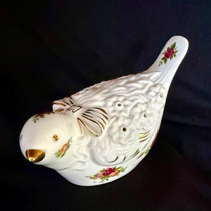 May include: A white porcelain bird figurine featuring a textured floral pattern on its back, a gold-painted beak, and delicate rose motifs. The bird has a stylized bow detail on its neck with gold trim accents and small holes across the back.