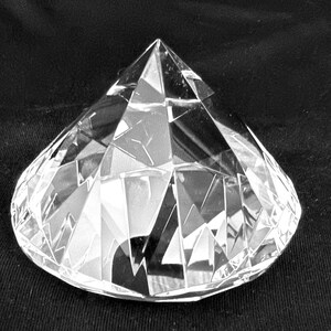 May include: A clear, faceted glass paperweight shaped like a large diamond. The object features numerous triangular and trapezoidal cut surfaces that reflect light, creating a geometric pattern across its transparent, colorless body.