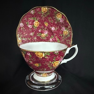 May include: A porcelain teacup and saucer set featuring a deep burgundy background with a repeating pattern of yellow and pink roses. The teacup has a white interior and a gold-trimmed scalloped rim, while the saucer displays a matching floral design.
