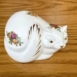 May include: A white porcelain cat figurine in a curled position, decorated with colorful floral patterns of pink and yellow roses. The cat features gold-painted accents on its side, ears, eyes, and nose, creating a detailed and reflective finish.