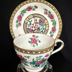 May include: A white porcelain teacup and saucer set featuring a hand-painted floral pattern with pink peonies, green leaves, and blue accents. The rims are decorated with a gold Greek key border, and the center of the saucer displays a detailed tree motif.