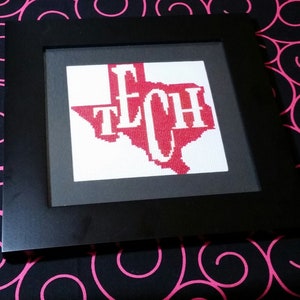 Könnte beinhalten: Ein gerahmtes Kreuzstichbild mit dem Umriss von Texas in Rot und dem Wort „TECH“ in Weiß. Der schwarze quadratische Rahmen steht im Kontrast zum weißen und roten Design, vor einem schwarzen Hintergrund mit rosa Wirbeln.