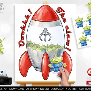 May include: A printable digital art sheet featuring a rocket-shaped claw machine filled with green three-eyed aliens. The text reads Ooohhh! The claw! in red letters. Includes a separate sheet of individual alien cutouts for assembly.