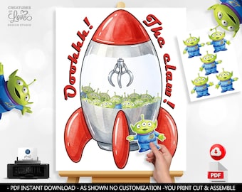 May include: A printable digital art sheet featuring a rocket-shaped claw machine filled with green three-eyed aliens. The text reads Ooohhh! The claw! in red letters. Includes a separate sheet of individual alien cutouts for assembly.