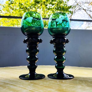 Puede incluir: Dos copas de vino de vidrio verde con un tallo y base verde oscuro. El cuenco de cada copa tiene un diseño de hojas grabado en el vidrio. Las copas se exhiben sobre una superficie marrón claro con un fondo verde borroso.