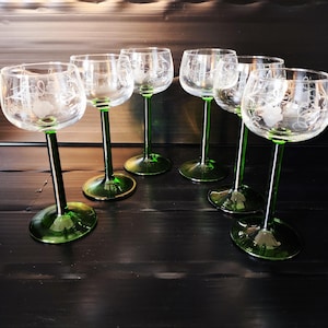 May include: A set of six clear glass wine glasses with etched floral patterns on the bowls, featuring long, slender stems and circular bases in a deep emerald green color, arranged in a semi-circle on a dark, horizontal surface.